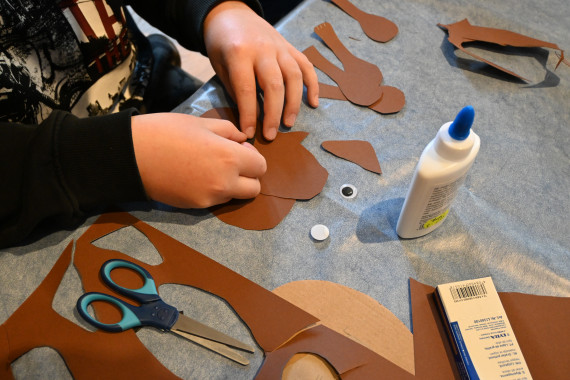 Tisch mit Bastelmaterialien (Tonpapier, Schere, Kleber); Man sieht die Hände eines Jungen, der daraus einen Hampelmann bastelt.
