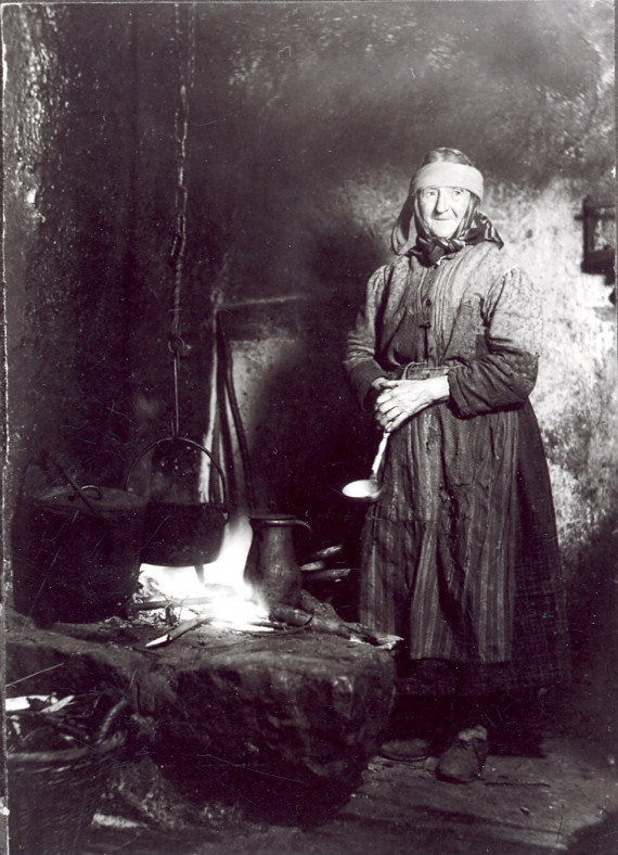 Schwarz-weißes Standporträt der Tagelöhnerin Theresia Stauder, um 1910, in ihrer Küche. Sie trägt ein langes Kleid mit langen Ärmeln, eine Schürze und ein Kopftuch. Die Kleidung ist alt und mit diversen Flicken repariert. In den gefalteten Händen hält sie eine Schöpfkelle. Neben ihr befindet sich eine Herdstelle mit offenem Feuer, über dem ein Kessel hängt. Die Wand hinter ihr ist schwarz vor Ruß. 