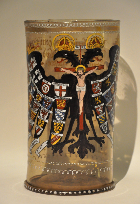 Ein großes, zylindrisches Glas aus dem 16. Jahrhundert, verziert mit Emaille. In der Mitte in schwarz ein großer doppelköpfiger, bekrönter Adler mit ausgebreiteten Flügeln. In der Mitte des Körpers der gekreuzigte Jesus. Auf den Flügeln Wappen ausgewählter Stände.