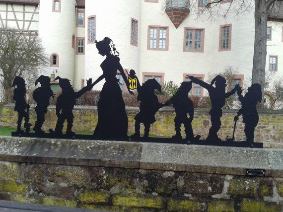 Foto einer zweidimensionalen Skulptur aus Blech.  Die Skulptur besteht aus schwarzem Metall und zeigt die Silhouetten von sieben Zwergen in unterschiedlichen Körperhaltungen und zwischen ihnen eine große Frau mit langem Kleid, Schleier und Krone (Schneewittchen). Der Zwerg rechts neben Schneewittchen hält eine gelbe Laterne in der Hand. Die Skulptur steht auf einer Mauer am Schlossplatz. Im Hintergrund erkennt man das Lohrer Schloss.. 