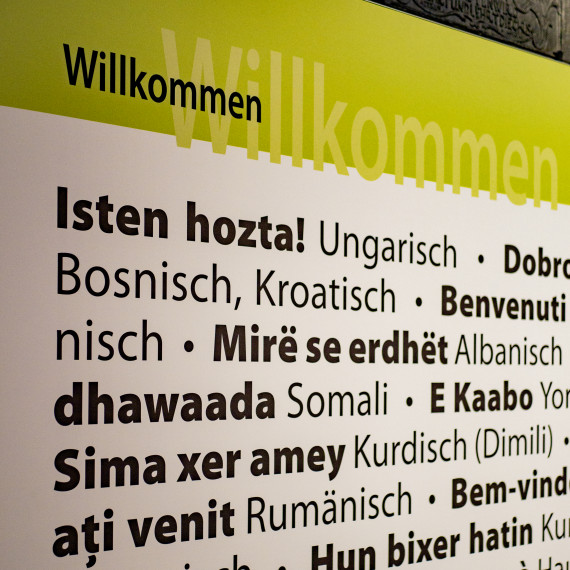 Detailansicht einer Texttafel der Sonderausstellung &quot;Woher | Wohin - eine Ausstellung vom Ankommen und Weggehen&quot;. Darauf zu sehen ist das Wort &quot;Willkommen&quot; in zahlreichen verschiedenen Sprachen. 
